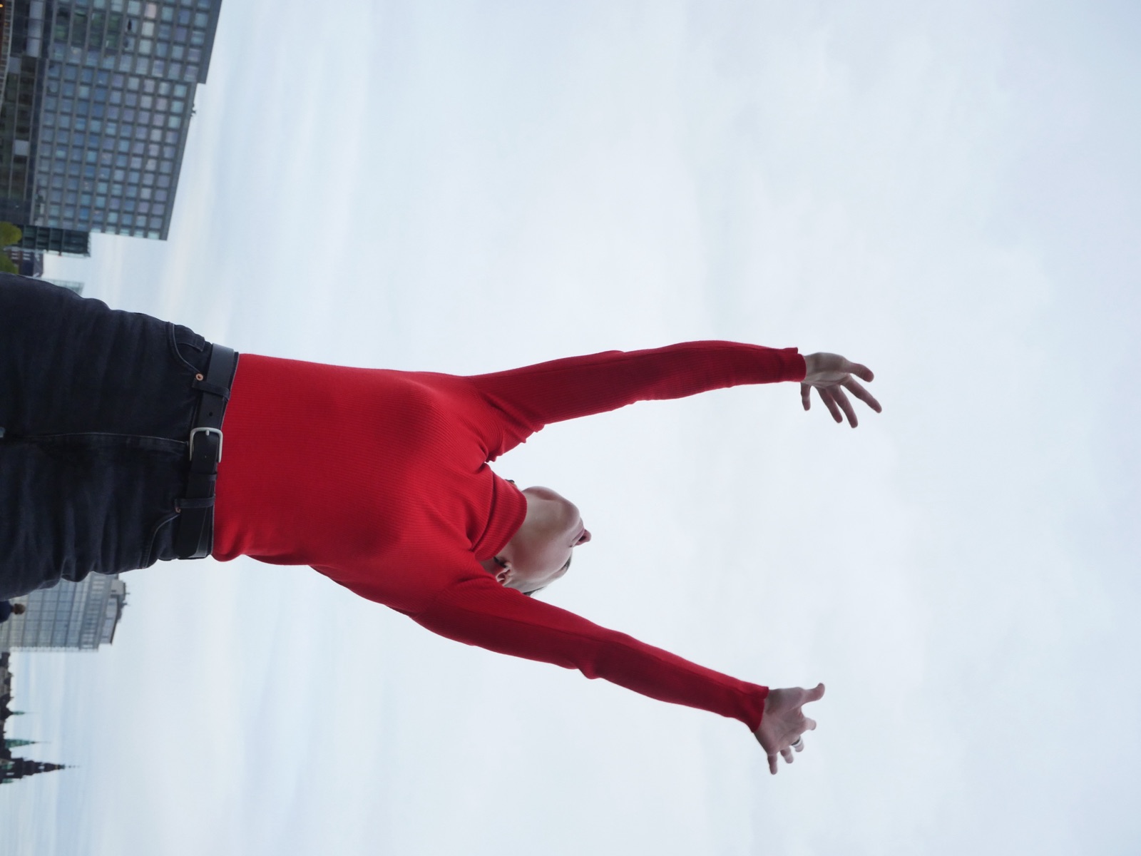Julia in a red top, arms flung open to a pale sky above a city skyline